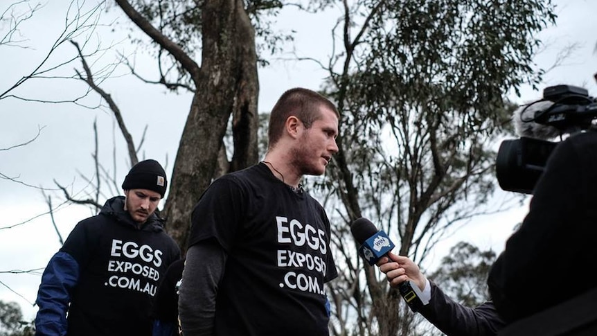 A man wearing a protest t-shirt stands in front of media and speaks into a microphone.