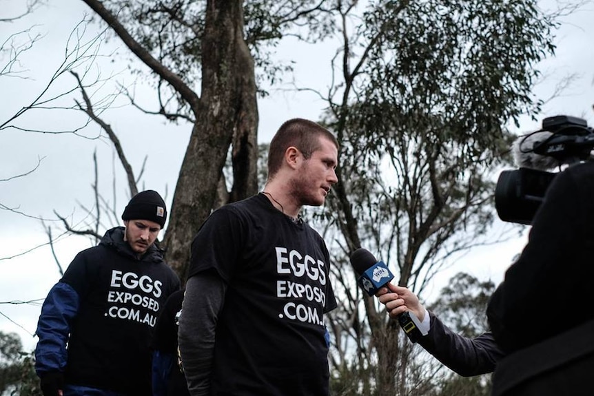 A man wearing a protest t-shirt stands in front of media and speaks into a microphone.