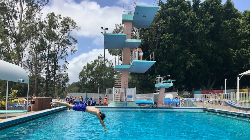 A child dives off a springboard into a pool with a high diving board at the far edge. 