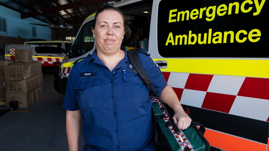 Tess wearing a dark blue paramedic uniform, holding a large case of equipment, standing next to an ambulance.