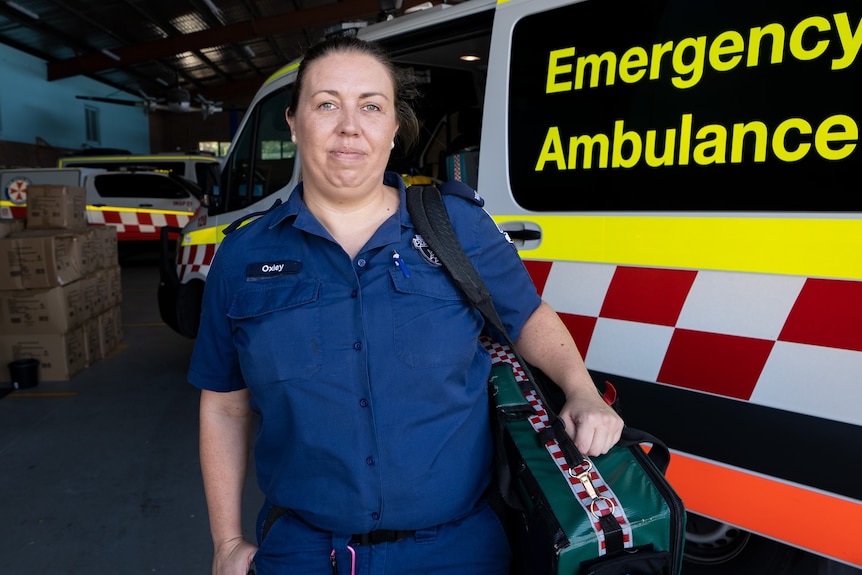 Tess wearing a dark blue paramedic uniform, holding a large case of equipment, standing next to an ambulance.