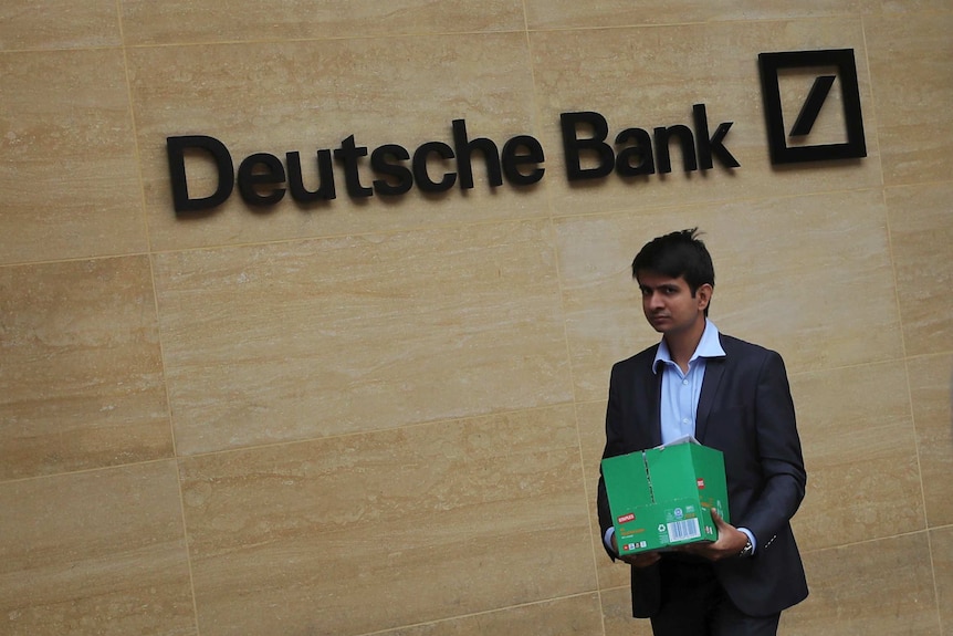 A man in a suit carrying a green box walks in front of a sandstone wall that carries the Deutsche Bank logo in London.