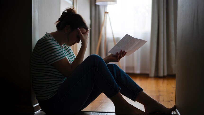 A woman sits holding a piece of paper with her head in her other hand, looking stressed. 