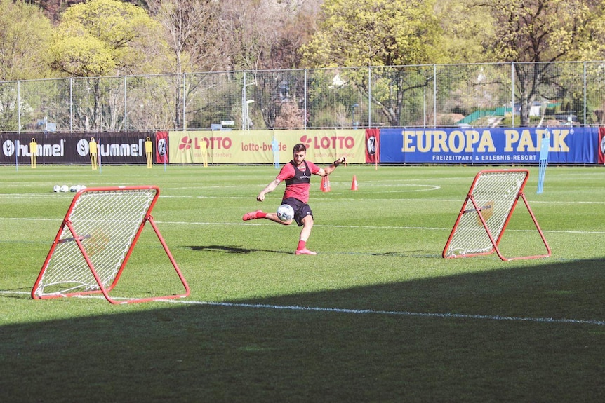 A man in a red shirt is pictured mid-kick on a soccer field with trees surrounding it.