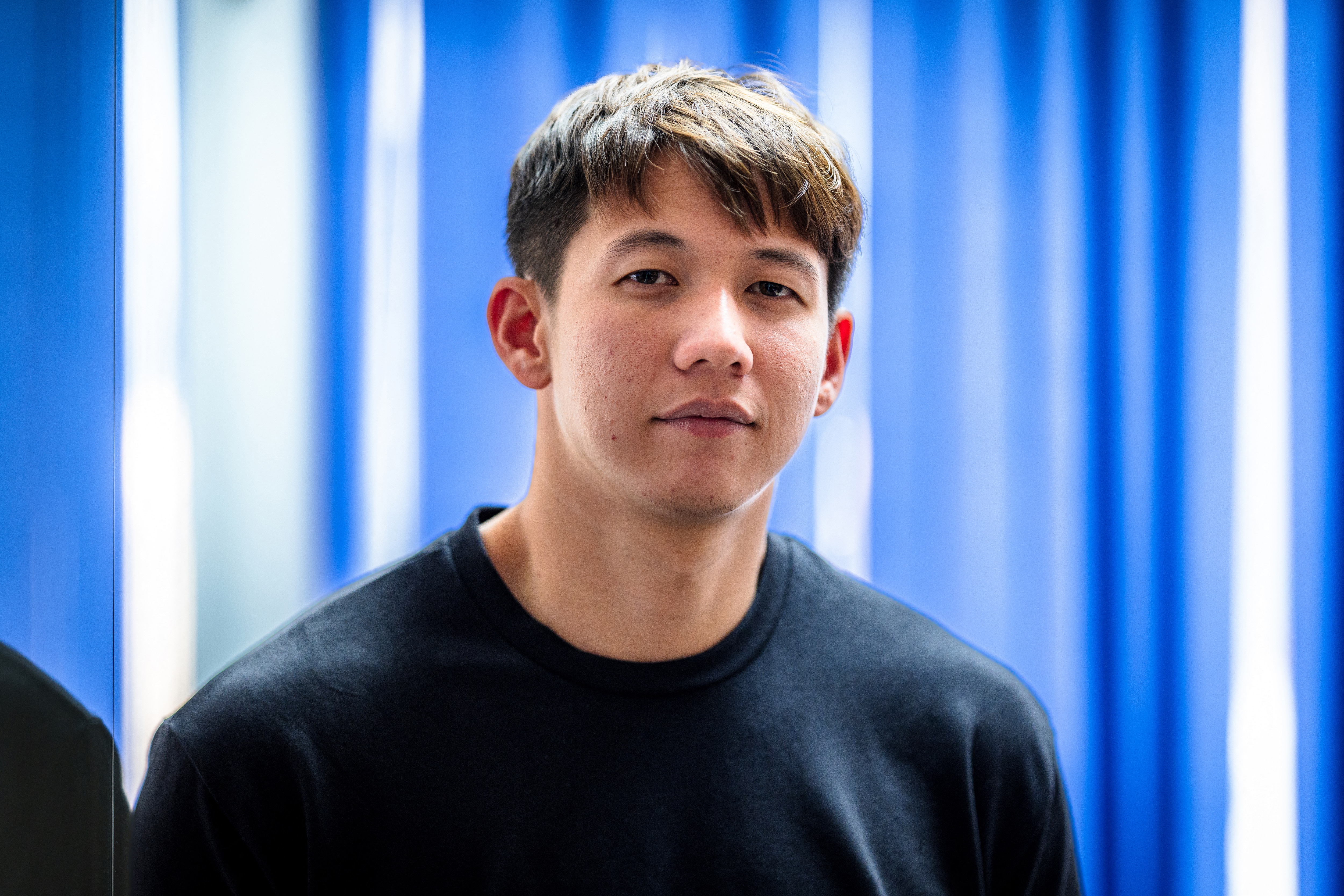 A man with short brown hair looks neutrally at the camera wearing a dark blue t-shirt in front of a blue background