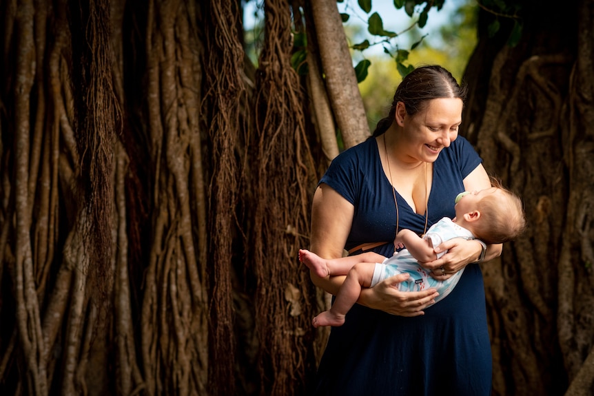  A mother wearing a navy dress carries a baby girl in a forrest.