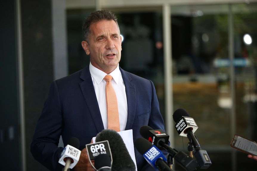 A man in a blue suit and light pink tie speaks to media 