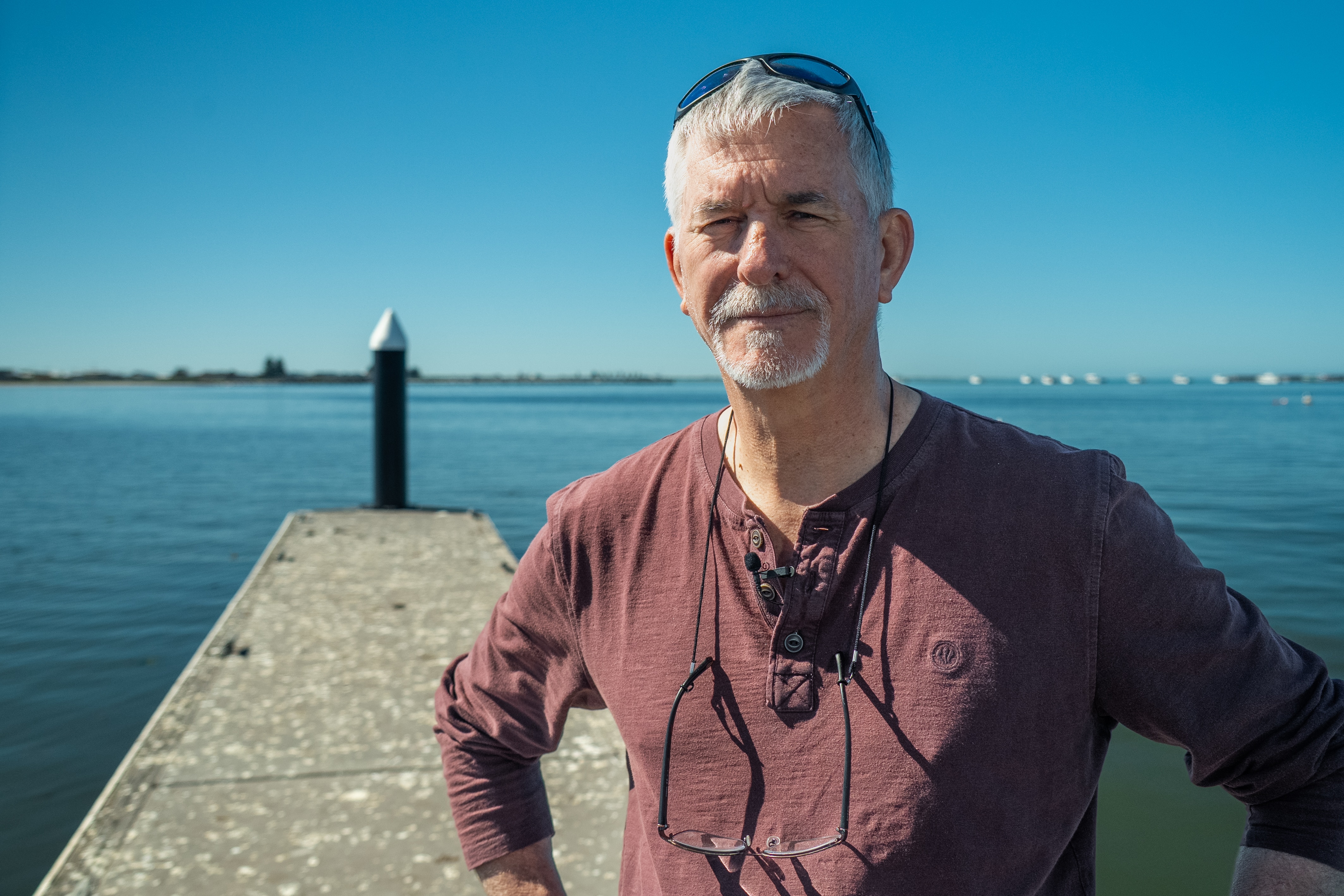 A man with grey hair looks at camera, standing on the pontoon of a coastal town on a sunny day.