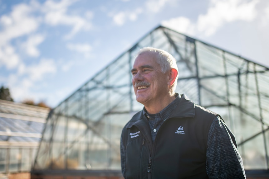 A gentleman with a mustache and white hair smiles outside transparent greenhouses with black frames under a partly cloudy sky