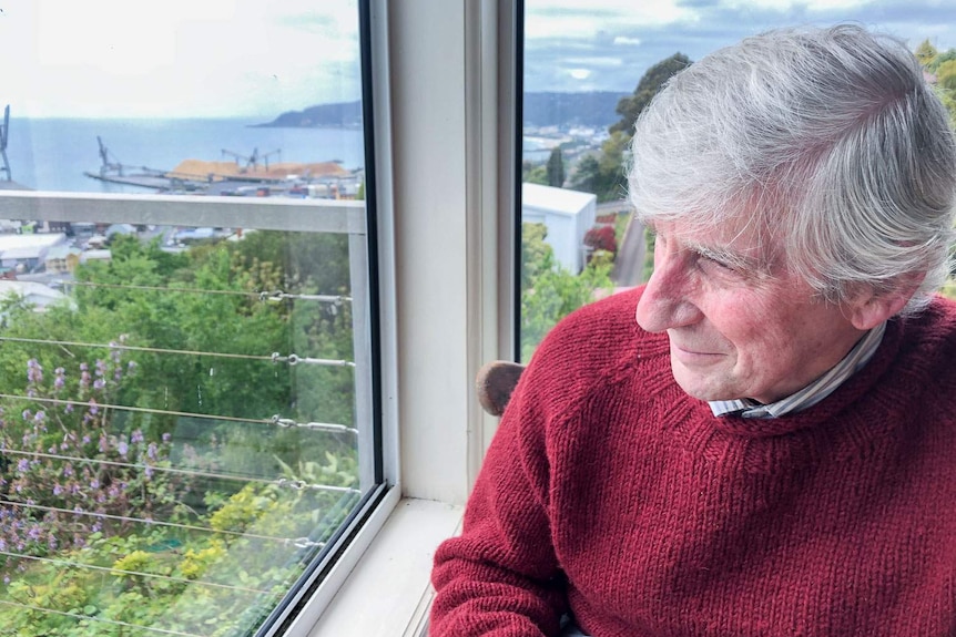 An older man gazes out window against a backdrop of Burnie and the port cranes.