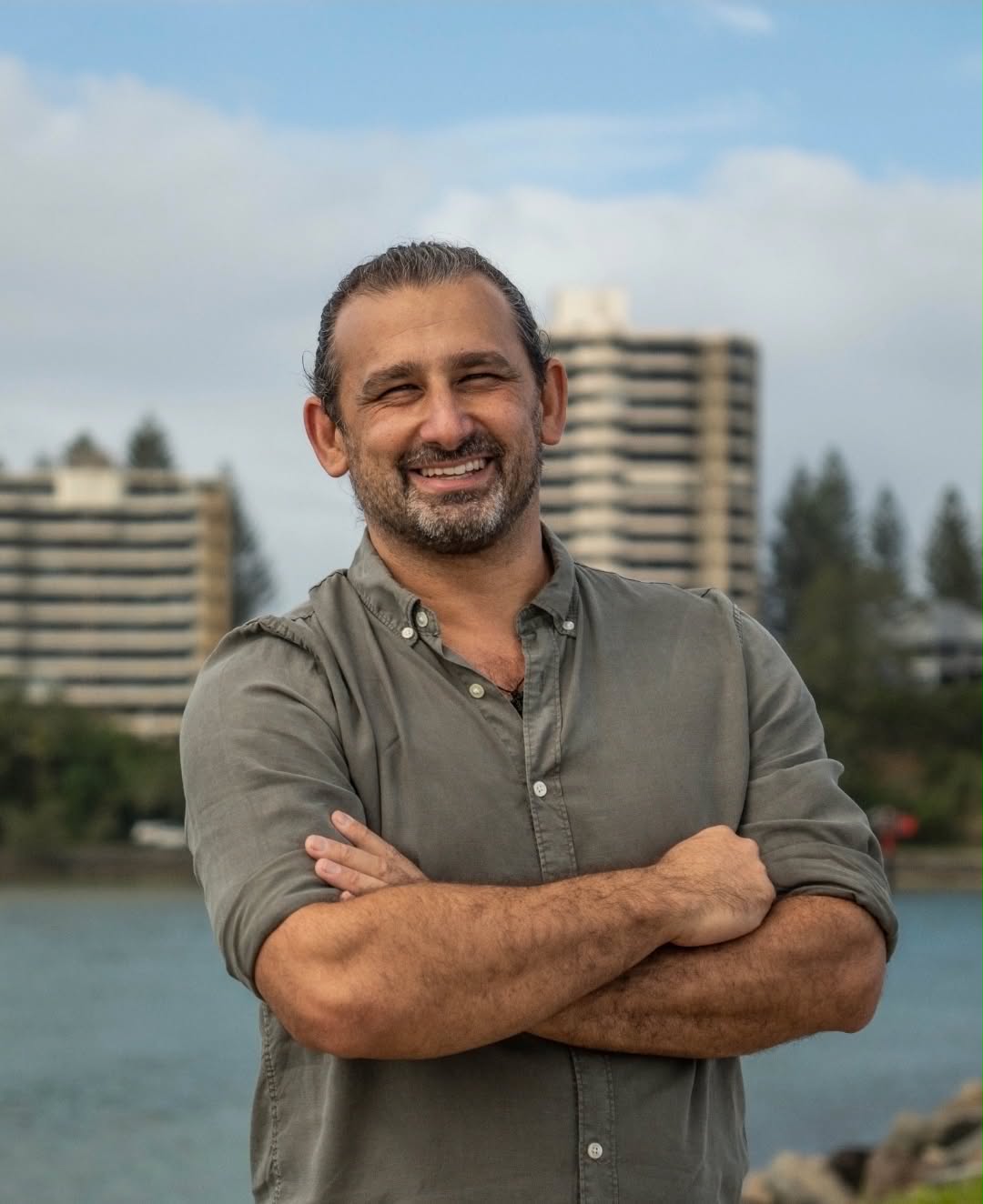 A middle aged man smiling at the camera, arms crossed, apartment buildings and houses in the background