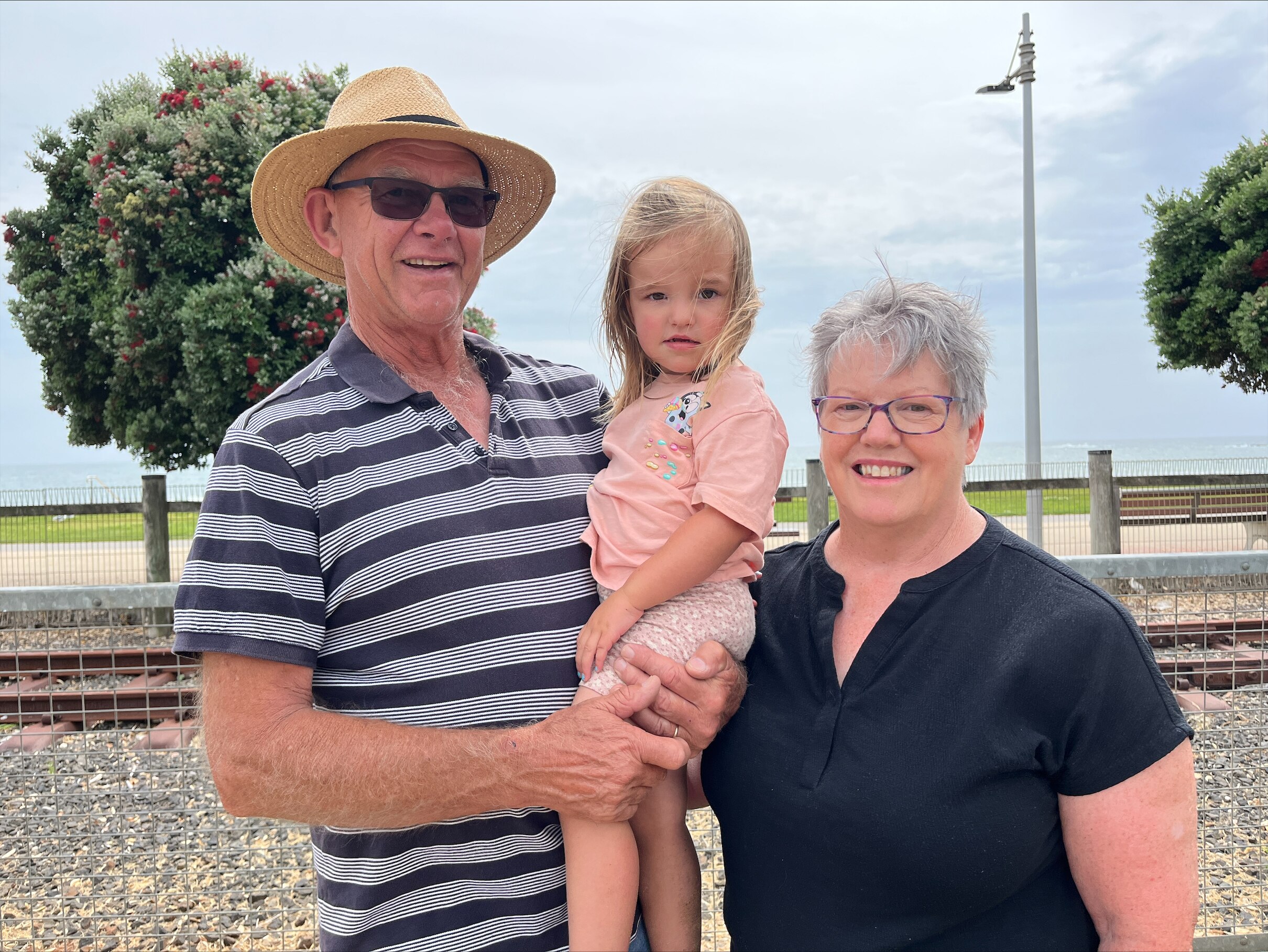 A woman a black shirt, a man wearing a white and blue striped shirt, holding a young child stand in front of a tree smiling.