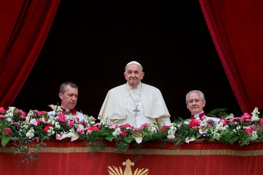Pope Francis stands on a balcony with two men either side of him and pink and white flowers in front of him.