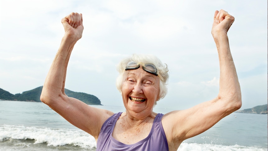 An older woman in purple bathers and goggles holds her fists high on beach