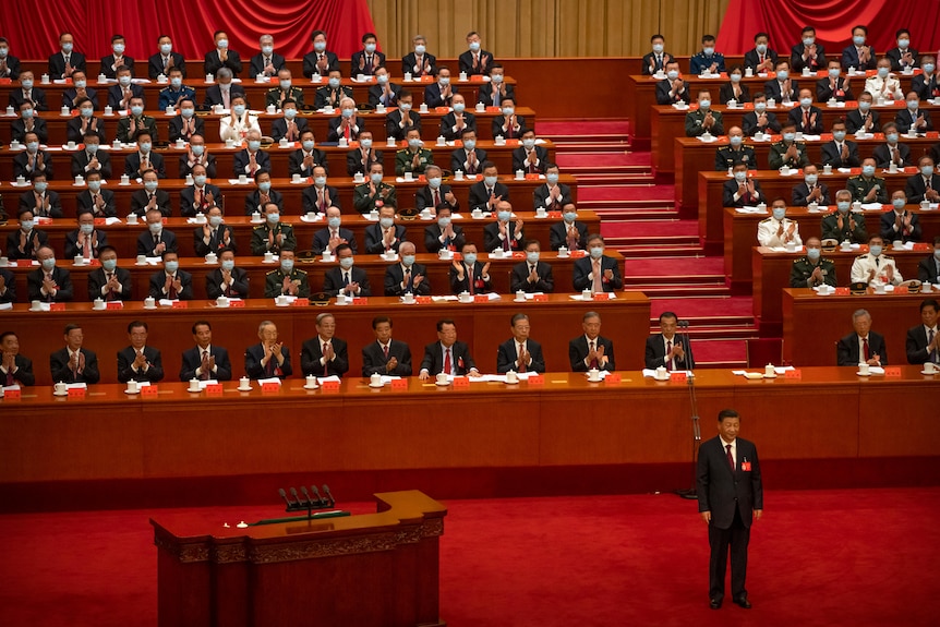 Rows of suited delegates sit behind a man who stands on a red carpeted floor.