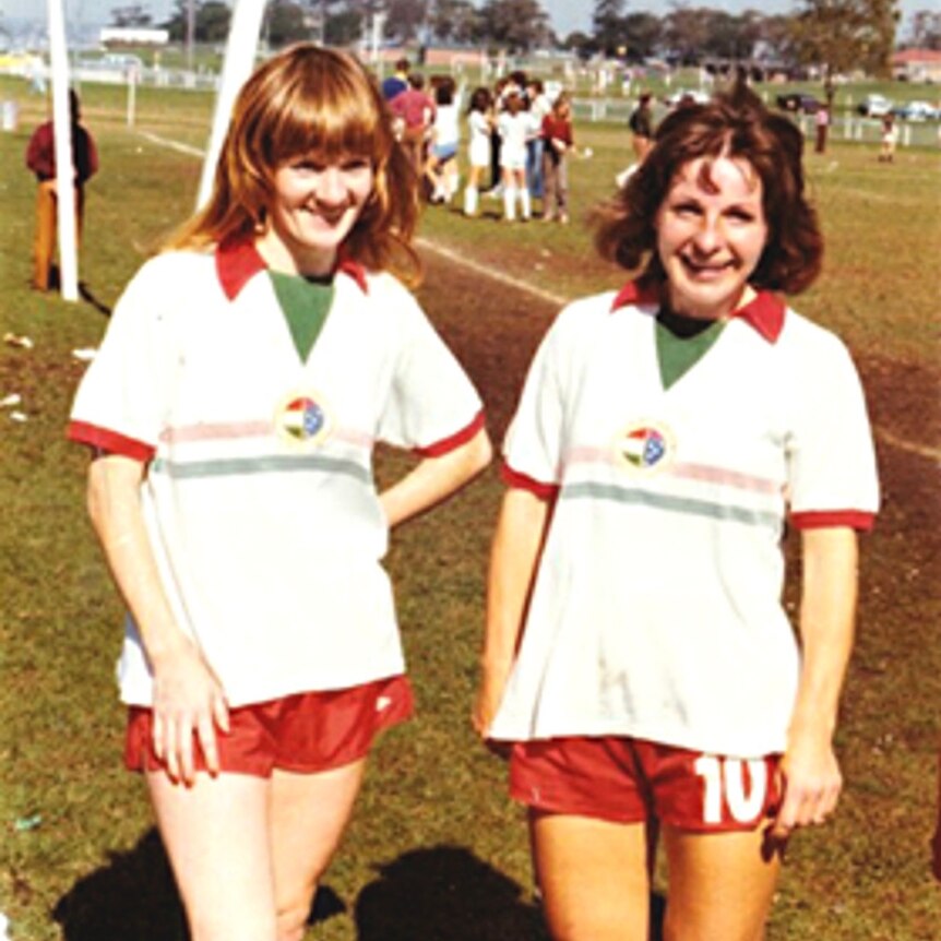 Two women soccer players wearing white jerseys with red shorts and green trim standing beside a grass field smiling