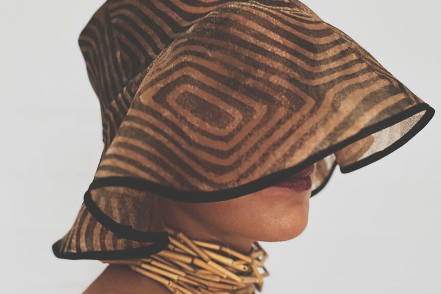 A model wears a hat and woven necklace, the hat obscuring all the face but the model's lips.