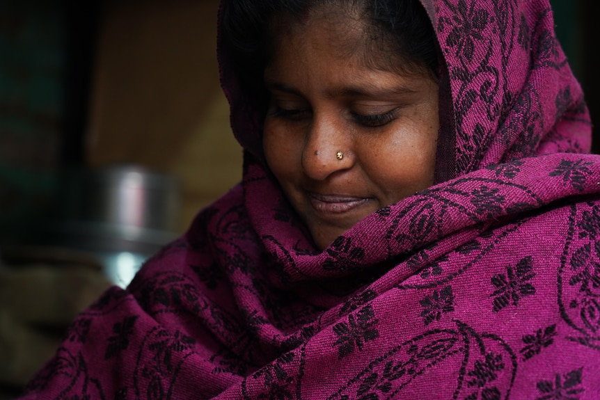 A woman wearing a floral patterned purple-maroon headscarf gives a subtle smile while glancing down