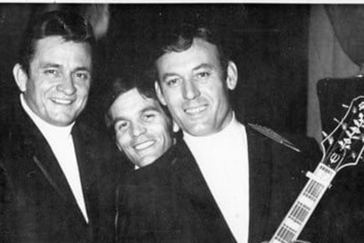 A black and white photo of three men in suits, one holding a Gibson guitar.