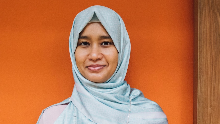 Raidah is shown wearing a light blue hijab, with an orange background behind her.