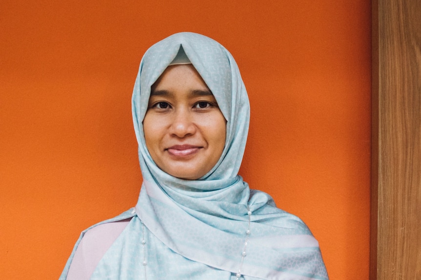 Raidah is shown wearing a light blue hijab, with an orange background behind her.