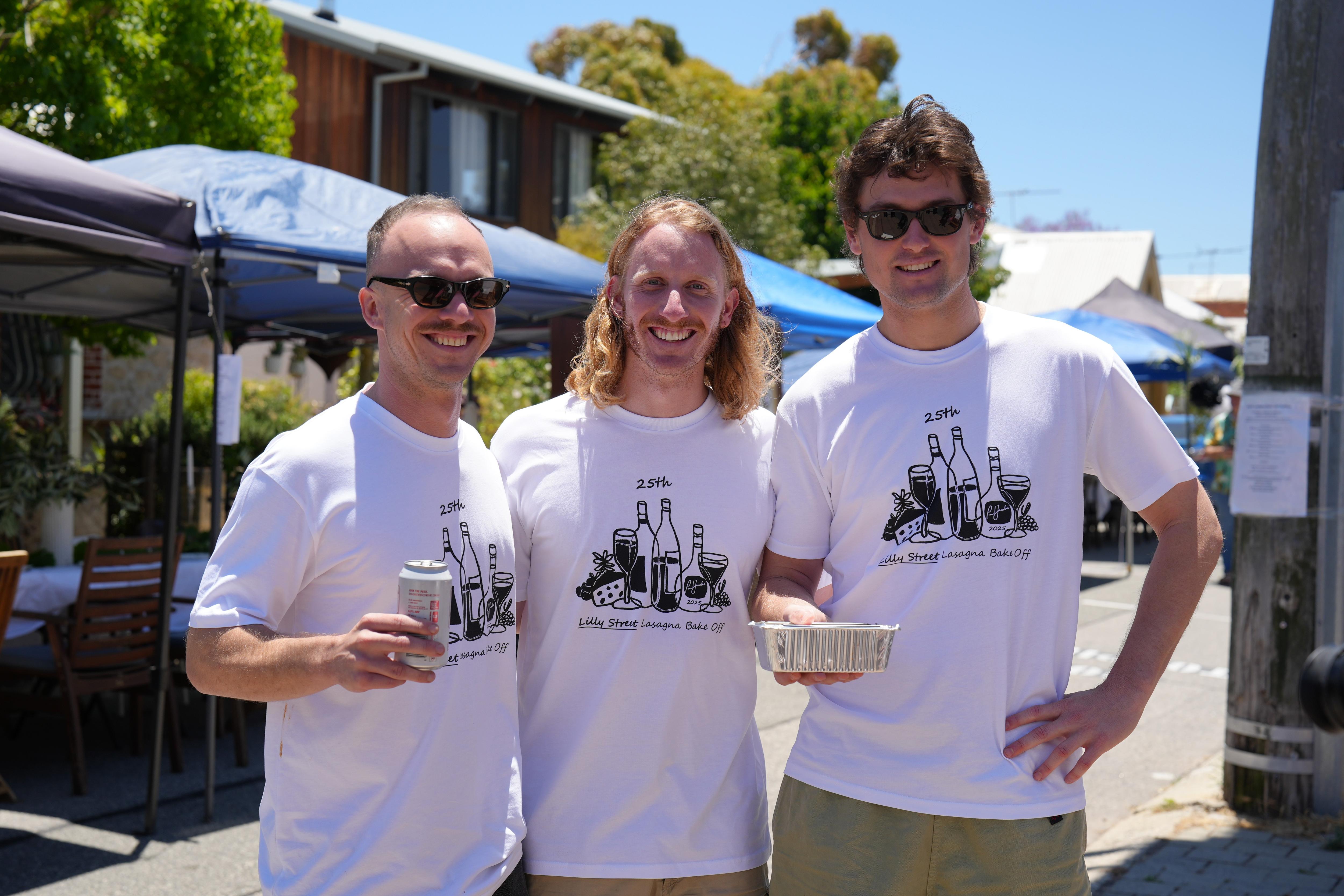 Tres hombres con camisetas blancas sostienen una bandeja de papel de aluminio con lasaña. 