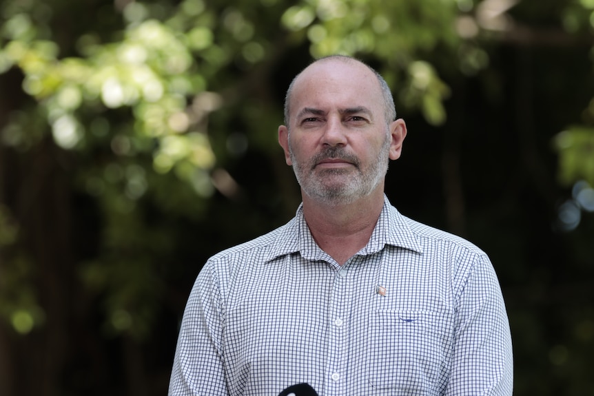 A man in a checkered collared shirt, standing outside with greenery in the background.