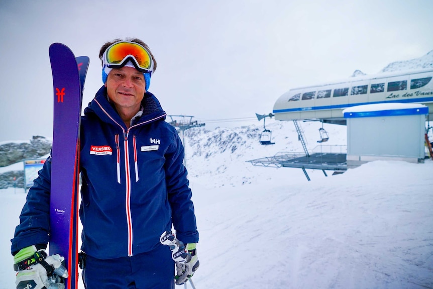 A man in a blue ski suit, goggles and gloves holding skis in the snow next to a chair lift