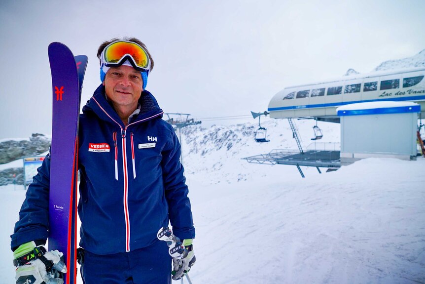 A man in a blue ski suit, goggles and gloves holding skis in the snow next to a chair lift