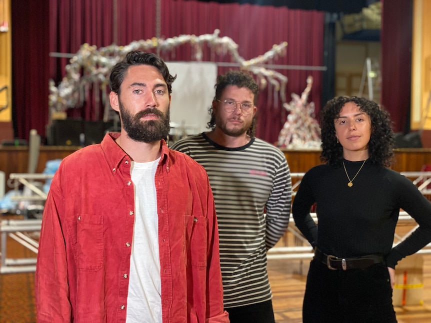 A man with a red shirt and beard stands next to another man in a striped shirt and a woman with curly hair