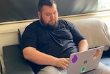 A man sits on a couch and looks at a laptop on his lap.