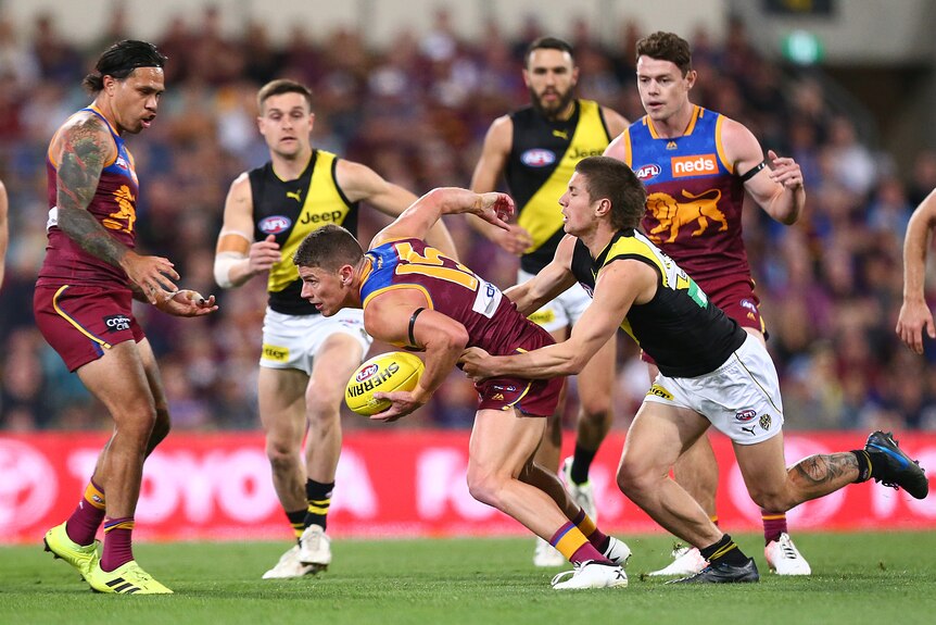 With a pack of players around the play, a Richmond player tackles the Brisbane Lions player holding the ball from behind