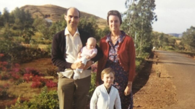 A family in the 1960s, standing by the side of a road, with trees in the background.