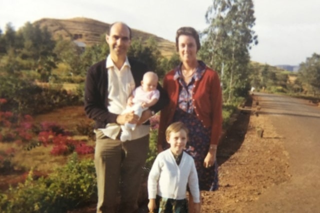 A family in the 1960s, standing by the side of a road, with trees in the background.