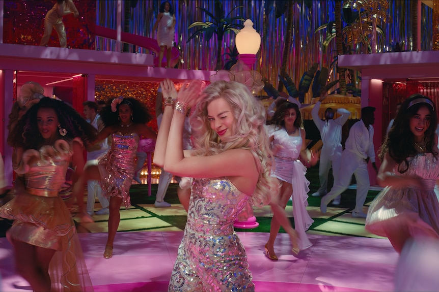 A blonde white woman in a silver sequined jumpsuit dances on a pink dancefloor surrounded by women in elaborate dresses.