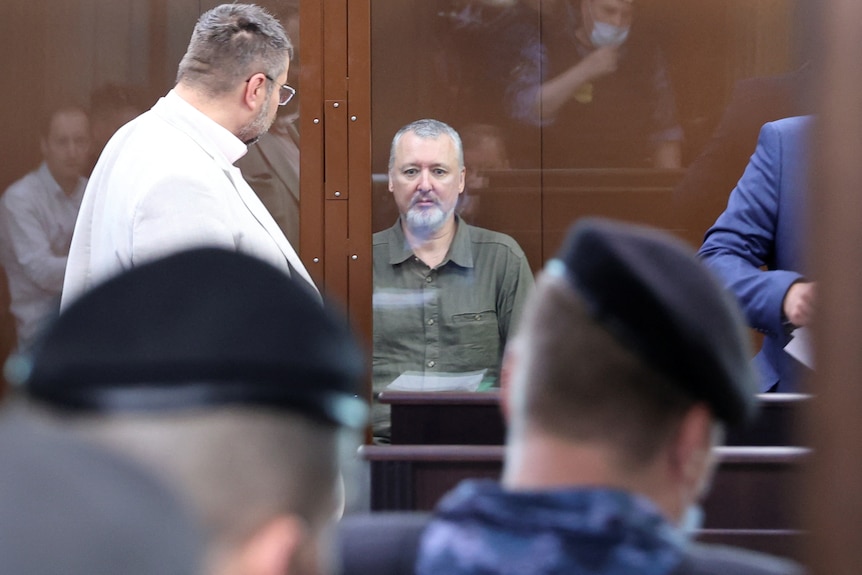 A grey haired man looking pale and confused in a glass cage 