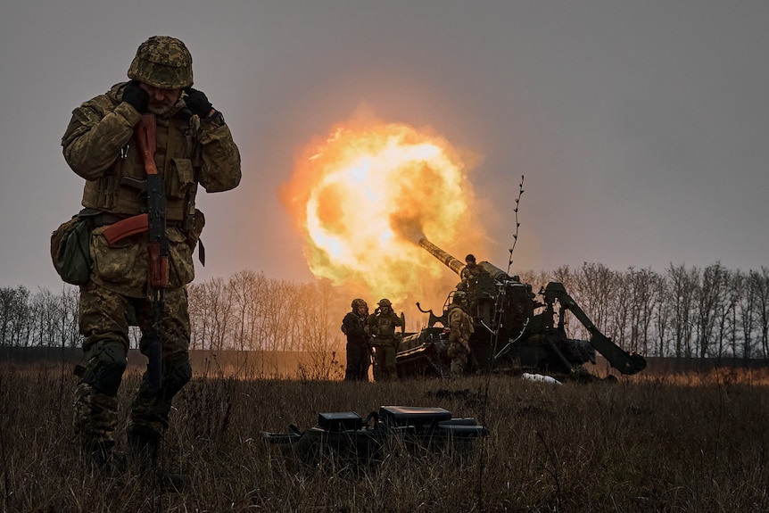 On the left a Ukrainian soldier with his fingers in his ears as other soldiers are firing an artillery system behind him.