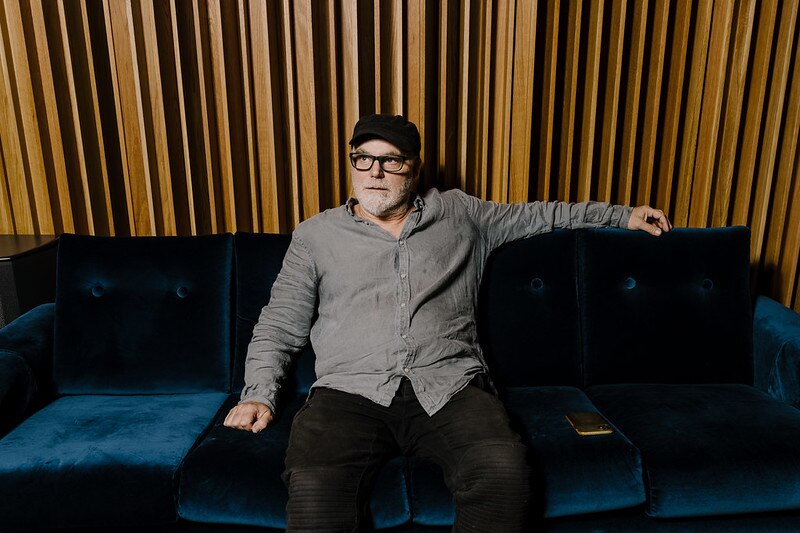A bearded man with glasses and cap sits on a blue velvet couch