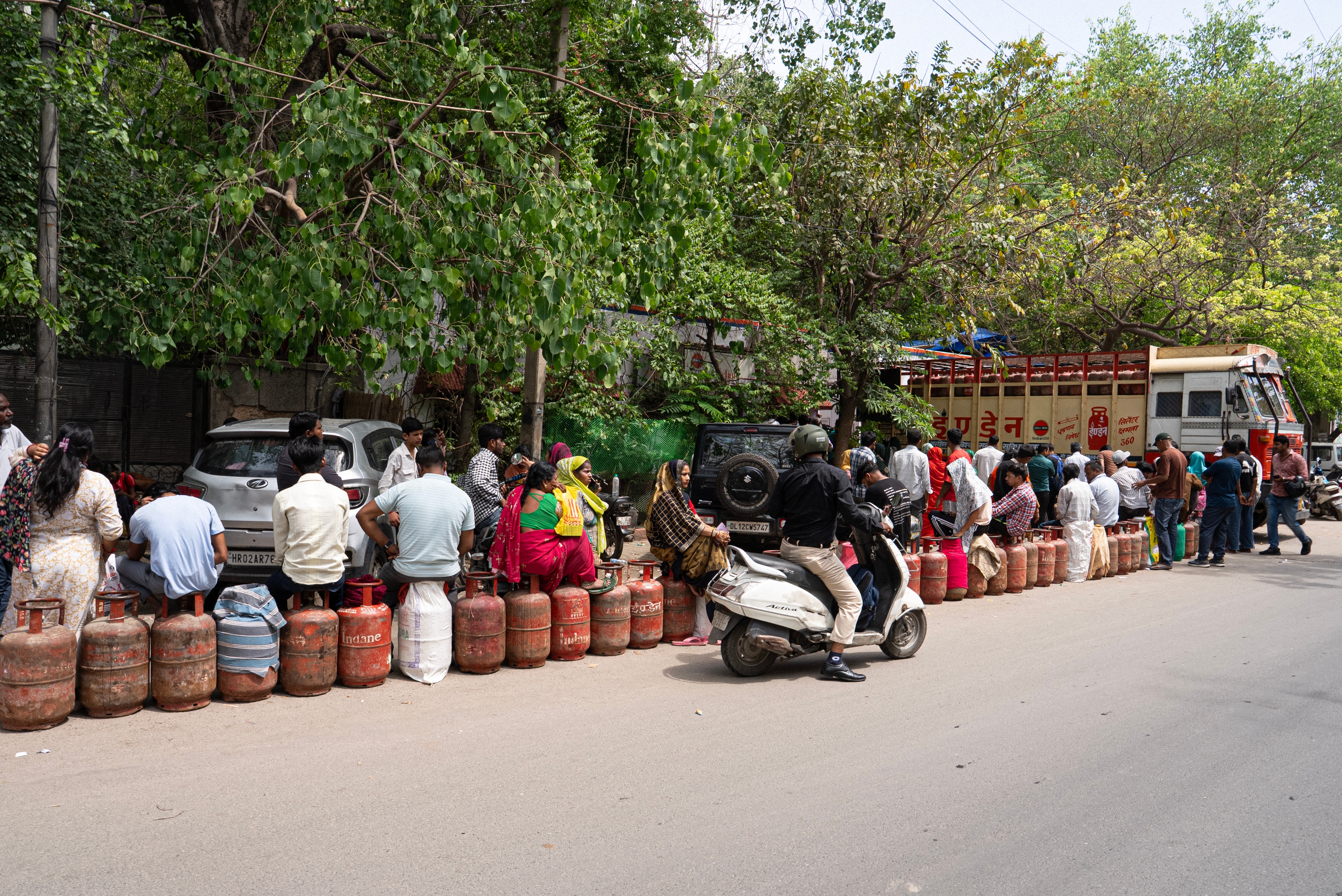 A group of people sit waiting in line in Delhi on gas cylinders, a truck parked in front, trees beside the road.