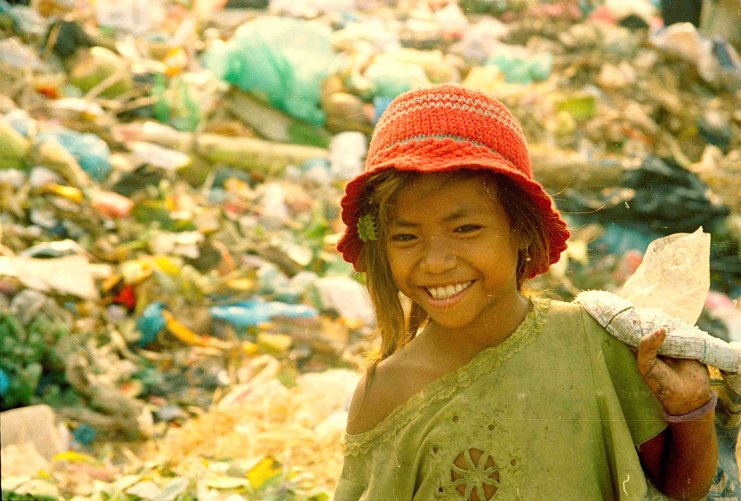 A girl, wearing dirty clothes, smiles as she walks past piles of rubbish
