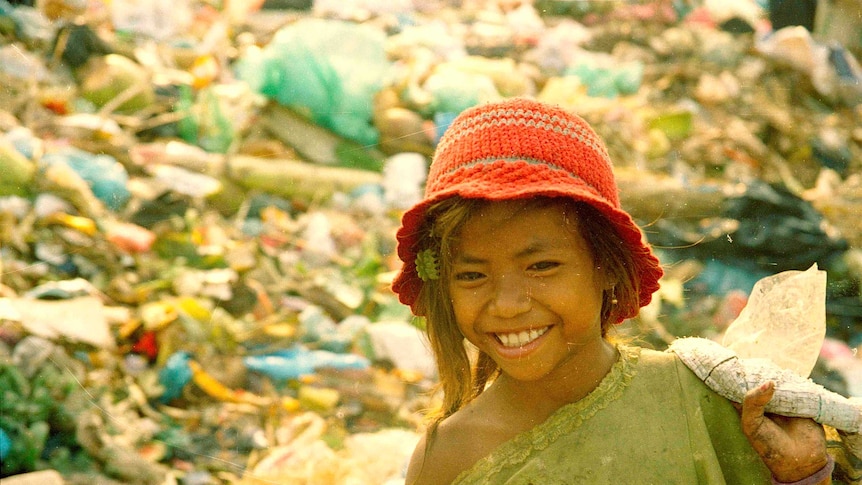 A girl, wearing dirty clothes, smiles as she walks past piles of rubbish