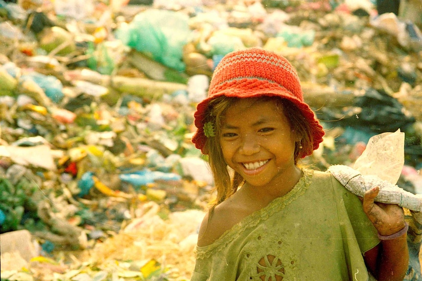 A girl, wearing dirty clothes, smiles as she walks past piles of rubbish