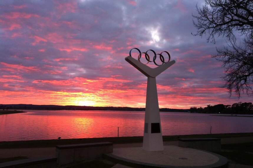 An Olympic Rings statue next to a lake at sunset