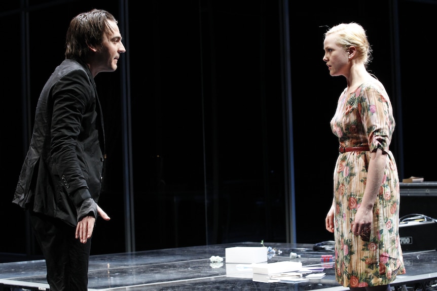 White man with dark hair wears all black suit and stands onstage before white woman with blonde hair and floral dress.