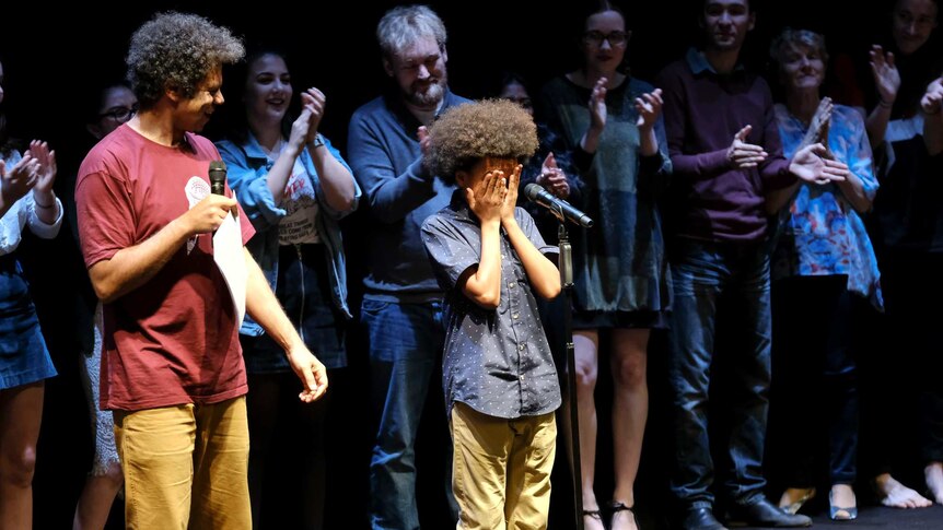Young boy has won slam poetry competition in Coffs Harbour, walking across stage while other competitors cheer in background.