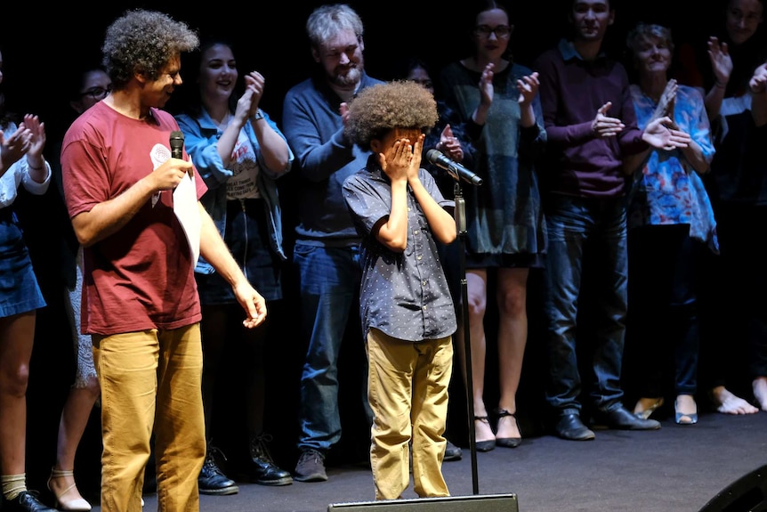 Young boy has won slam poetry competition in Coffs Harbour, walking across stage while other competitors cheer in background.