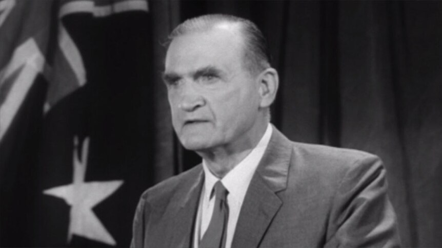 A black and white photo of a man, standing in front of an Australian flag
