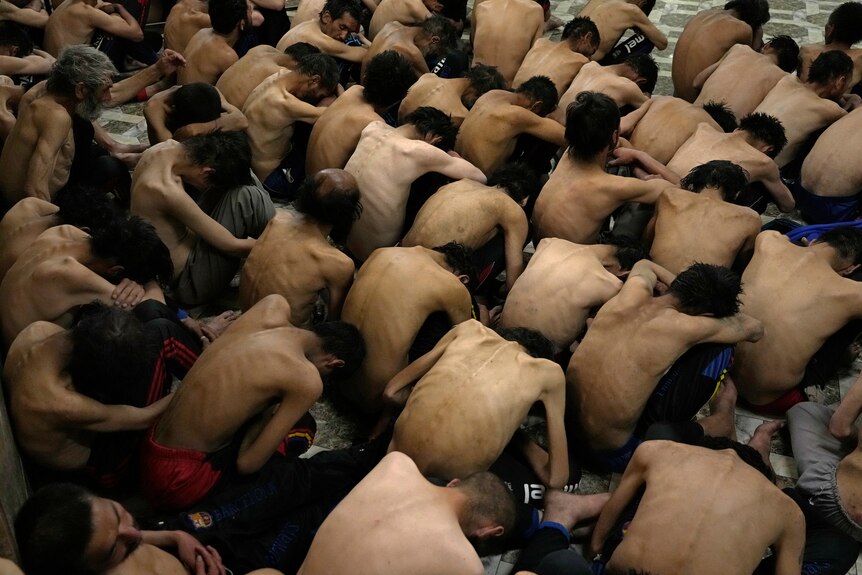 Rows of skinny shirtless men sit hunched on the floor.