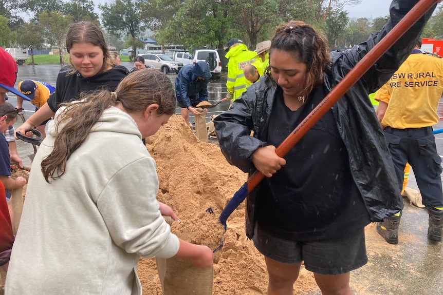 Two girls stand next to a pile of sand using a spade to fill bags.