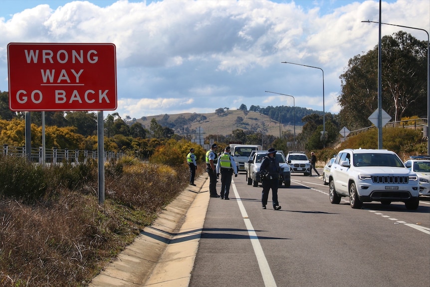 A red sign says wrong way go back in the foreground and in the background police check cars on a highway.